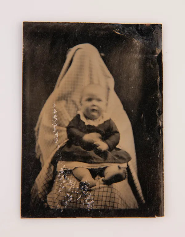 [Tintype portrait of a baby held in the lap of a person concealed by cloth].